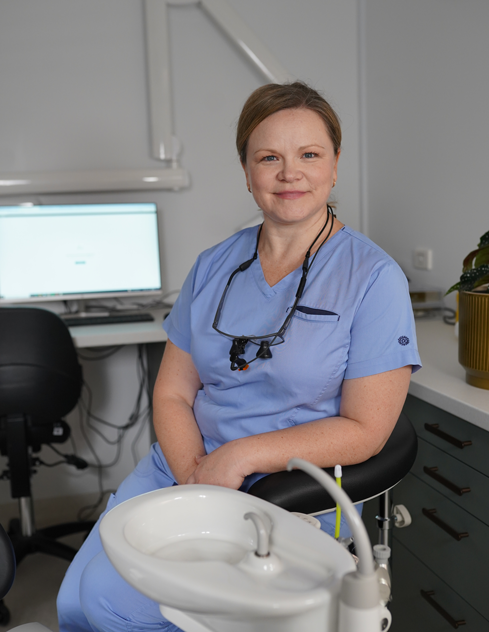 Park Orchards Dental | Smiling dentist in blue scrubs seated beside a dental chair in a treatment room